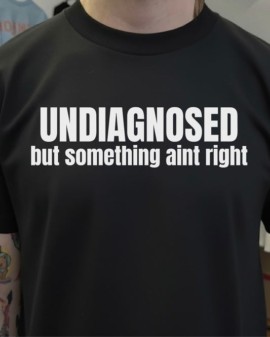 Black t-shirt with white text 'UNDIAGNOSED but something aint right' worn by a person.